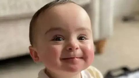 Close up of the smiling face of the late nine-month-old Genevieve Meehan. She has brown eyes and is wearing a lemon and white outfit.