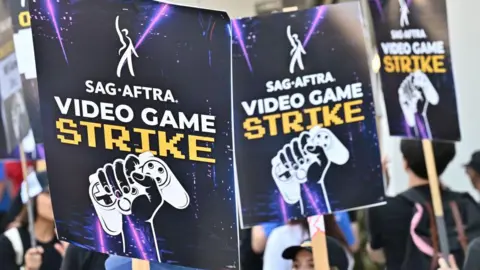 Getty Images Three placards being held in the air with a picture of a fist clenched around a gaming controller and the text "Sag-Aftra on strike!". 