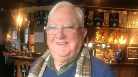 Marie Wilson Image of a man in pub. He has white hair, glasses and a tartan scarf. He is smiling.