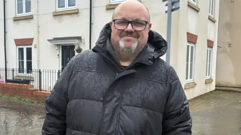 A middle aged man wearing a black puffer coat and black glasses. He's smiling, stood in front of a white house. 