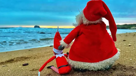 Pat Christie A toy santa and elf photographed from behind sitting on a sandy beach with blue waves visible in the background.