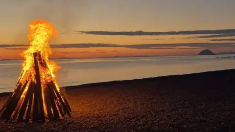 Colin Shearer Orange flames emerging from a beacon made from wood. It is on a beach in front of a yellow and blue sunset.