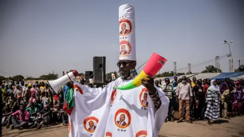 A man, who is wearing a hat and tunic bearing the letters "CDP", holds a megaphone and horn