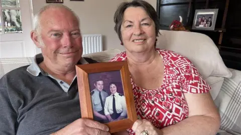 Ed and Madge Ryan, sitting on a sofa, holding a picture frame with them in their police uniform. Ed has on a grey top and Madge, a red and white top. They are smiling at the camera. 