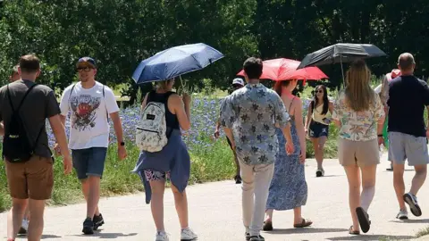 Getty Images About seven people walking through a park on a concrete path with trees and flowers beside them. Three are holding umbrellas to shield them from the sun. Most are in shorts and T-shirts.