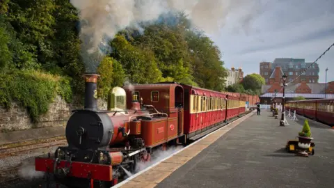 A steam train at Douglas Station. The engine is brown and there is smoke flowing out of the chimney. Red and cream coloured carriages stretch back behind the engine alongside the platform.