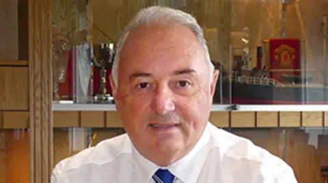 John Laird sitting behind a desk in a trophy room. He has short white hair, and is wearing a white shirt and blue tie.