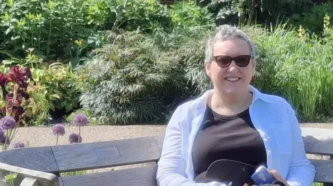 PA Media Tracy Tomlinson with short grey hair wearing a pale blue cotton shirt over a black t-shirt and sunglasses. She is sitting on a wooden garden bench in a garden. She is smiling.