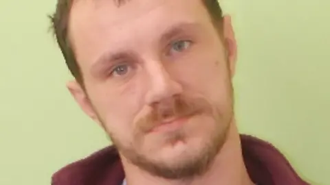 Guernsey Police Adrian Dodd - A man with brown hair and a brown beard and moustache leaning his head to his right looking at the camera. he is wearing a burgundy jumper and a blue T-shirt. He is holding a board with his prisoner number on it and is standing in front of a green background. 