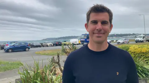 Tom Pinsard is smiling at the camera as he stands on a garden. Behind him is a road which has cars parked along it next to a harbour wall with the sea extending beyond. He has short brown hair and is wearing a navy crew cut jumper.