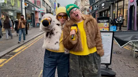 @lyndseyandlouise Louise and Lyndsey Scott are pictured standing on a busy street in Liverpool with shops lining both sides of the road. One wear a yellow woolly hat and the other wears a bright green hat, both are in sunglasses, fur jackets and bright yellow tops, posing and holding microphones. 