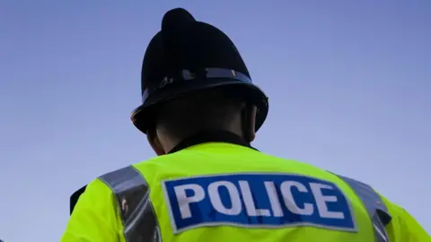 A policeman stands with his back to the camera. He is wearing a police helmet and a high-vis jacket. In the background is blue sky 