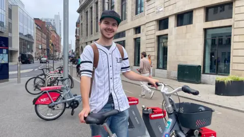 Erin McLone/BBC A man is stood beside an e-bike with one hand on the seat and another on the handlebars. He wears a white baseball style top with black buttons and stripes, a brown backpack over his shoulders, blue skinny jeans, a black cap. He is stood by the docking bay at the side of a road.