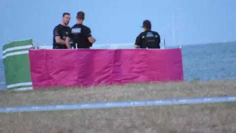 Lewis Adams/BBC Three police officers standing on the beach in West Mersea. They are surrounded by green, pink and white windbreaks. Blue and white tape - indicating a cordon is in place - is pictured in the foreground.