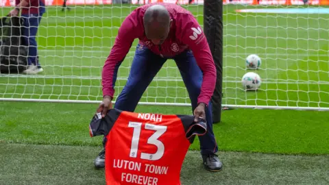 Luton Town FC A man holding a Luton Town football club top in memory of Noah, which he is placing by the ground behind the goal at Wembley Stadium. The shirt has Noah written on it and the number 13 and the words: "Luton Town Forever".
