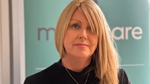 Teresa Cope, who has long blonde hair and is wearing a black top and a gold pendant necklace. She is sitting in front of a sign that says Manx Care on a light green background.