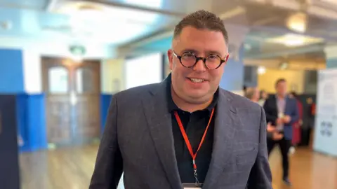 BBC Former fraudster Alex Wood is pictured at the Oxford Fraud Conference. He's wearing an orange lanyard, grey checked suit and round tortoiseshell glasses.