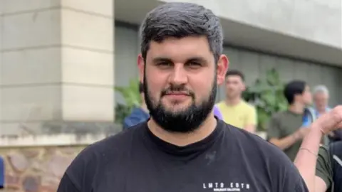 A man stood outside a hospital that has been evacuated. He is wearing a black t-shirt.