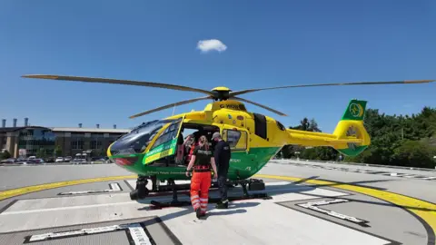 A yellow and green helicopter stationary on a helipad with a building in the background, a door open on the helicopter with one person standing inside and a white man in dark clothes and grey baseball cap and white woman in black t-shirt and orange trousers standing outside looking in