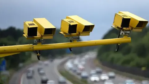 Three yellow roadside speed enforcement cameras mounted on a horizontal pole above a multi-lane highway, with traffic visible in the blurred background. The cameras are positioned to monitor vehicles traveling along the road.