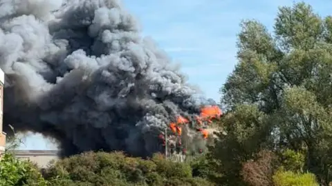 Hayley Curtis A green metal building is on fire. Large flames and thick black and grey smoke are coming from the building. There are several trees around it.