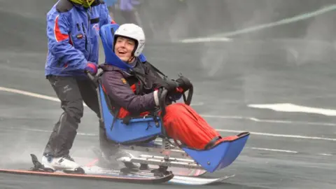 Kendal Snowsports Club A person who is disabled is skiing using an adaptive seat and ski. They are wearing purple and red waterproofs and are being pushed down by another skiier. The adaptive skiier is laughing and looks happy.