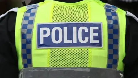 A police is standing with their back towards the camera. They are wearing a black jacket and a yellow hi-vis that says POLICE on the back.