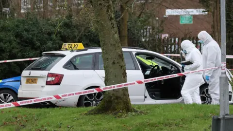 A white estate car with a taxi sign sits in a car park. There is a police cordon tied to a street sign. A marked police car is parked behind the taxi. There are a variety of trees and patches of grass at the perimeter. Two forensic officers in white body suits examine the taxi.
