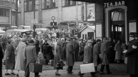 Crowds of people in overcoats with suitcases queue at Blackburn Railay station in a 1957 BBC documentary.