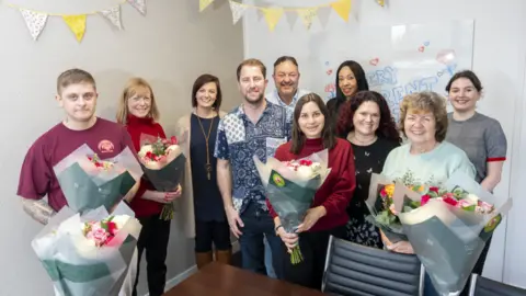 Swiis Foster Care A group of about ten people, men and women, all standing in a bare conference-style room with yellow bunting across the back walls. They are smiling to camera behind a desk and chair and several of them hold large bouquets of flowers.