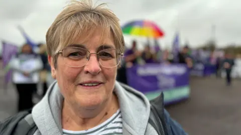 Maura McKenna standing with unison members outside Stormont. She has short blonde hair and is wearing light coloured framed glasses, a grey hoodie and a dark blue and black coat. You can see purple unison flags in the background and a rainbow umbrella.