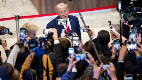 Reuters Donald Trump wearing a blue suit, white shirt and red tie stands in front of press holding camera phones and microphones up. 
