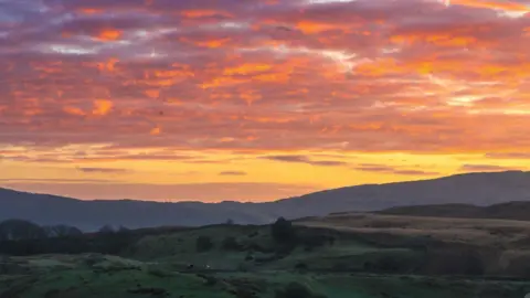 BBC WEATHER WATCHERS/ JUDE An orange and red glow across a cloudy sky just before sunrise. Trees and hills below have not yet been lit by the sun.