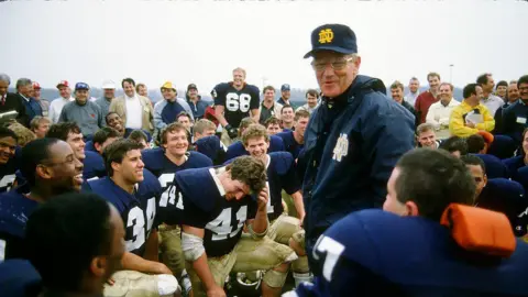 Lou Holtz standing at the centre of a group of kneeling players, and speaking. He's in a baseball cap and windbreaker with the Notre Dame logo 