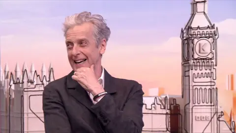 Peter Capaldi, dressed in a suit, smiling and turning to one side in TV studio
