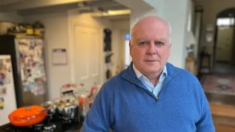 Paul Arnott, leader of East Devon District Council, in a blue shirt and blue zip-neck jumper in his kitchen