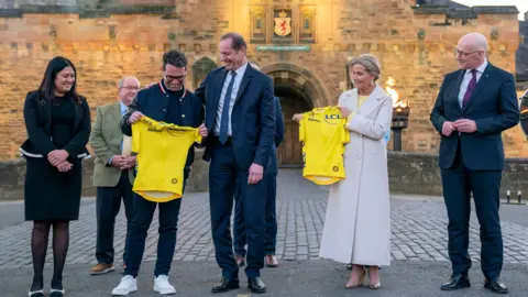 PA Media Lisa Nandy, Mark Cavendish, Christian Prudhomme, the Duchess of Edinburgh and John Swinney standing in a row outside the front gate of Edinburgh Castle. Cavendish is being handed a yellow Tour De France jersey by Prudhomme.