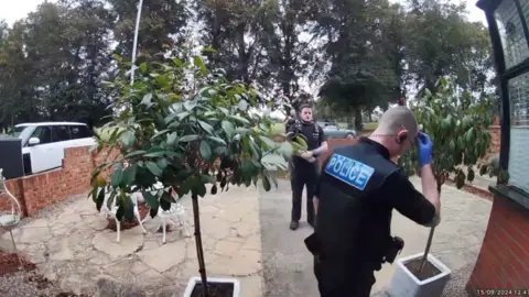 Police officers appear at the door of Maria Botterill's home in Northampton. At least three male agents are seen. They are standing in the driveway, which is closed off by a wall. There are large trees in the background.