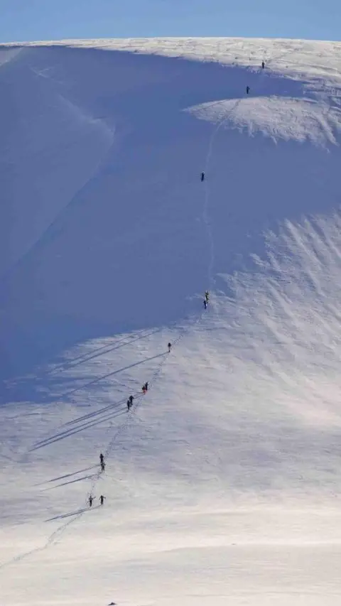 SAIS Southern Cairngorms Ski tourers walking up a snow covered slope at Glas Maol in the Southern Cairngorms. The skiers are walking in a line up a steep slope under a blue sky.