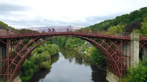 Robyn Furtado A wide angle of the red, metal Ironbridge, which stands above the River Severn. There are three cyclists riding over the bridge. On either side of the river are green trees and bushes, as well as a small white house on the banks of the river.