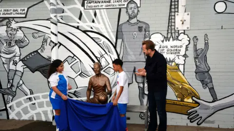 PA Media Two young football players can be seen pulling a blue sheet off the statue at its unveiling. Harry Kane is seen clapping to the right of them. They are standing in front of a black and white comic-style mural showing moments from Kane's career.