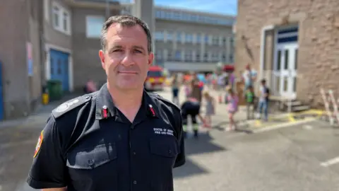 The image shows Paul Brown standing in the foreground wearing a dark fire and rescue service uniform, facing the camera in an outdoor courtyard or service area between two stone buildings. There are children behind him near a fire engine.