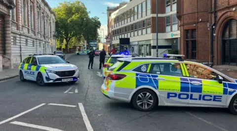 Police cars parked in the middle of a city centre street. It is daytime. Police officers are in the background.