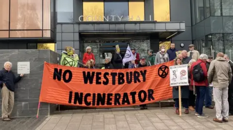 LDRS A group of people holding an orange sign which says "no Westbury incinerator" outside council offices.
