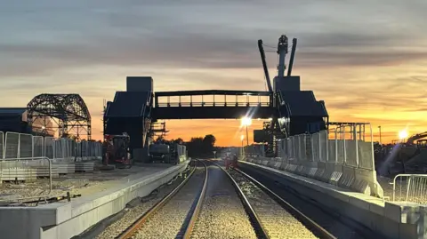 Weca A bridge crosses two train platforms, as seen from the centre of two rail tracks. The bridge is an outline, with the sun and orange-grey skies behind.