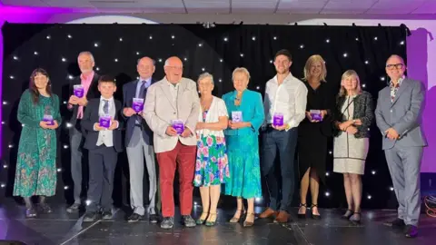 A group of people stand in a line on a stage, and are looking at the camera and smiling. They are holding purple awards in their hands and the background is a black backdrop with white lights shining on it.