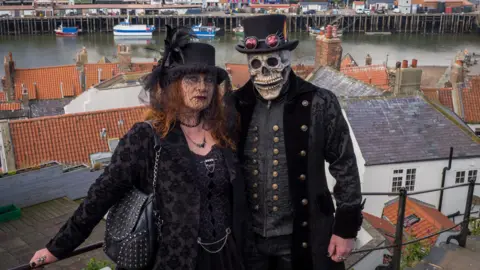 A man and a woman dressed up for the Whitby Goth Weekend. The woman is wearing a black jacket, black hat with lace veil and carries a leather handbag. The man has a black top hat and is wearing a black velvet jacket with golden buttons. His face is covered with a skull mask.