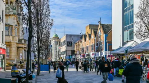 Getty A busy pedestrian Ilford high street lined with a mix of modern and older buildings, including a historic stone building on the left. People walk along the wide paved walkway, some carrying bags, while market stalls and shopfronts line the right side. Leafless trees and a bright blue sky complete the scene.