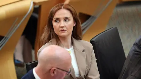 Getty Images Màiri McAllan wears a beige blazer and white top. She has long brown hair and looks off to the side while sitting next to John Swinney in the Scottish parliament