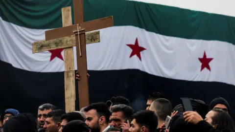 Christians march in Damascus, holding up two crosses, with a large Syrian flag in the background